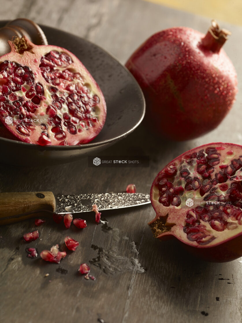 Pomegranate cut in half with one half in a bowl and the other on a wood table with seeds around the base