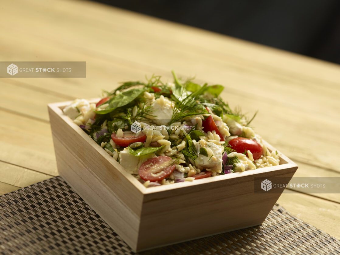 Orzo pasta salad with cherry tomatoes, spinach, feta, red onion and pine nuts in a wood catering bowl on a placemat