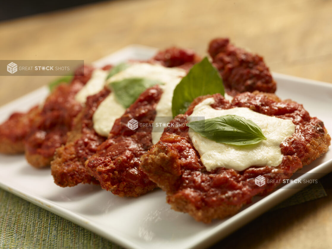 Pieces of chicken parmesan arranged a white ceramic platter, garnished with fresh basil, close up view