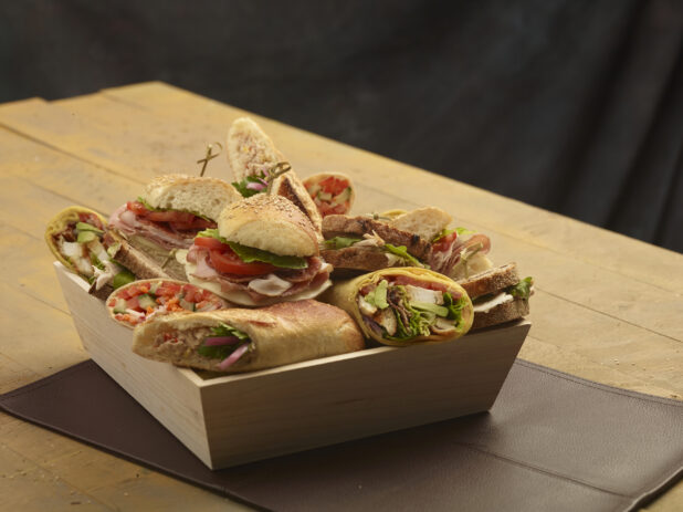 Assorted rustic sandwiches and wraps in a wood catering box on a wooden background