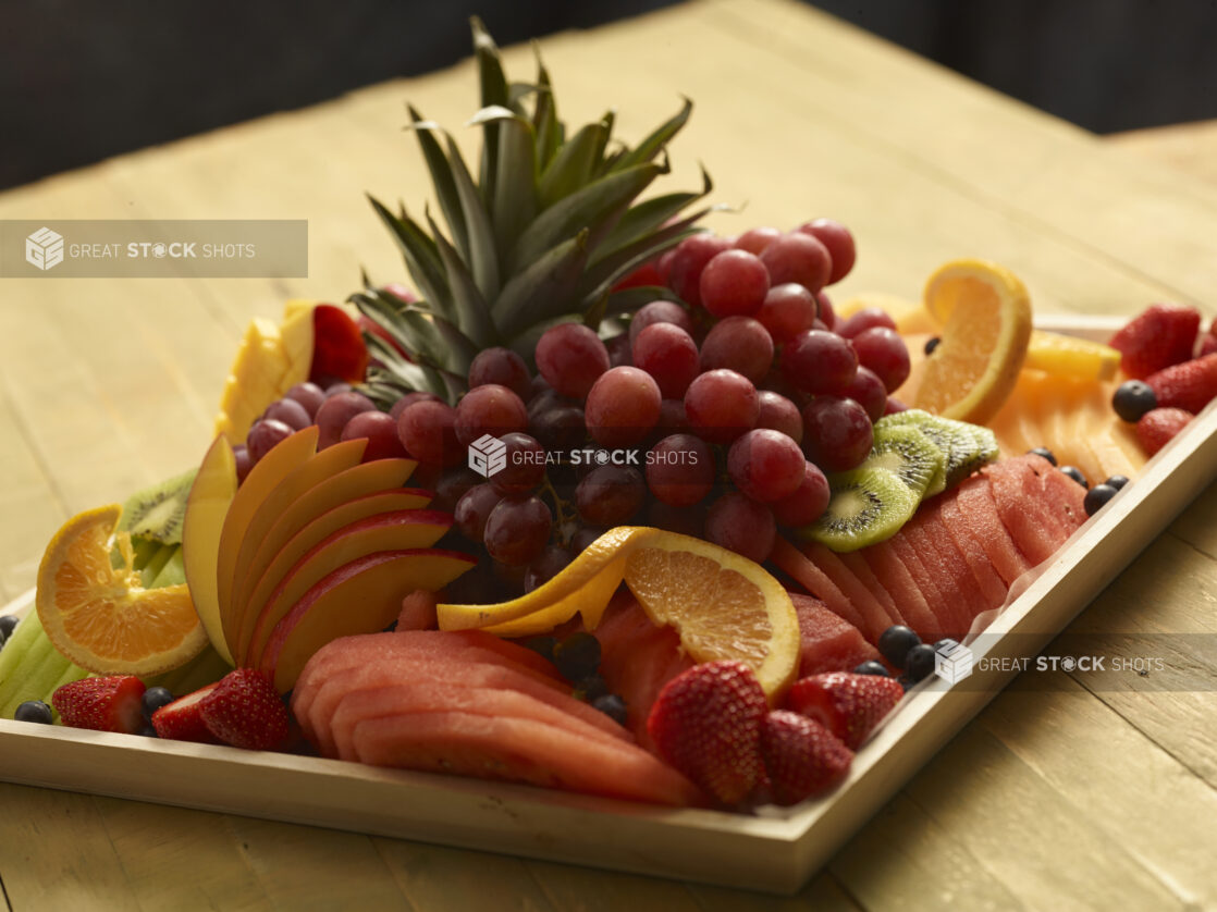 Fruit platter with grapes, watermelon, mango, strawberries, oranges, blueberries, melon, kiwi, cantaloupe on a wood catering tray on a wooden background