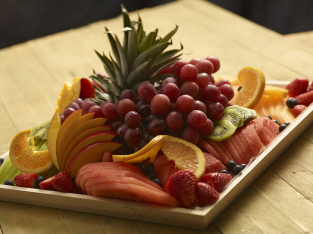 Fruit platter with grapes, watermelon, mango, strawberries, oranges, blueberries, melon, kiwi, cantaloupe on a wood catering tray on a wooden background