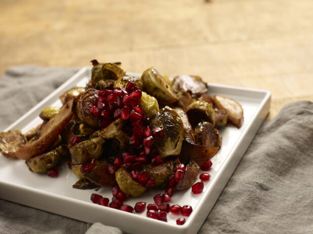 Roasted brussels sprout, potatoes and sweet potatoes garnished with fresh pomegranate seeds on a small square white ceramic plate