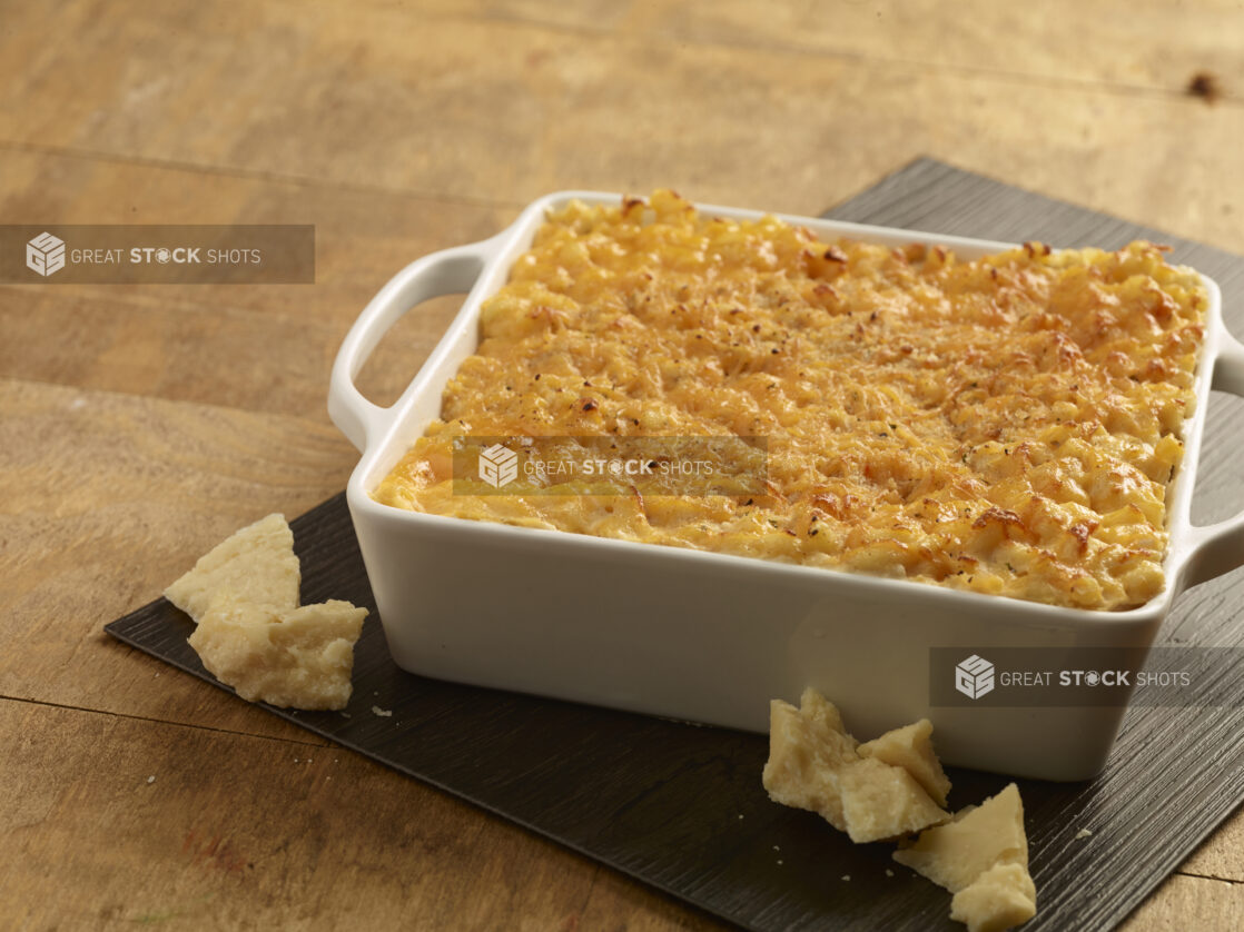 Baked macaroni and cheese in a white ceramic square dish with chunks of parmesan cheese in the foreground