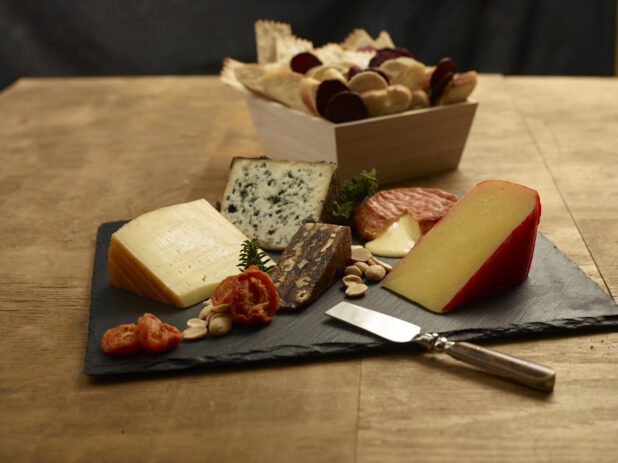 Cheese tray with various cheeses, sundried tomatoes and nuts on a slate platter with assorted breads and crackers in a wood catering box in the background
