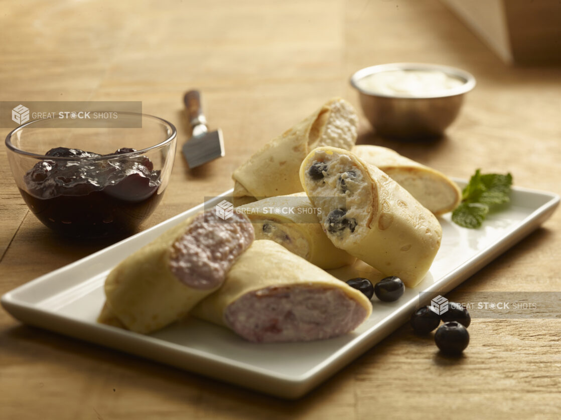 Blintzes (sweet crepes) garnished with fresh blueberries and mint on a rectangular white plate with jam in a small glass bowl and sour cream in a ramekin on a wooden background