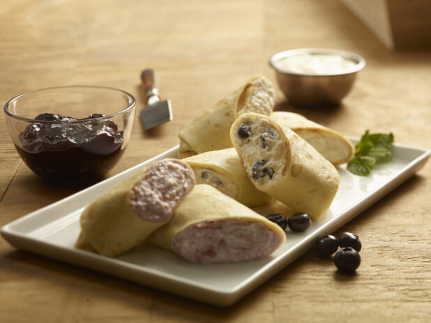 Blintzes (sweet crepes) garnished with fresh blueberries and mint on a rectangular white plate with jam in a small glass bowl and sour cream in a ramekin on a wooden background