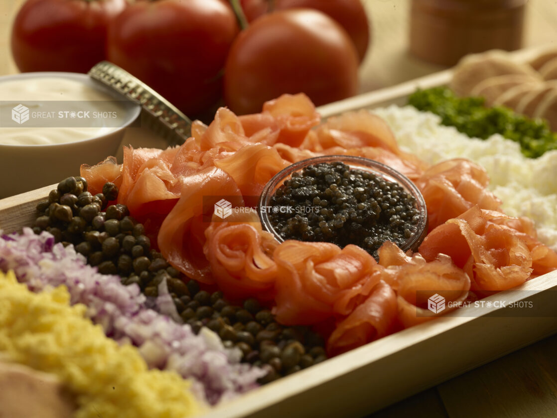 Close up view of a smoked salmon platter with red onions, capers, caviar, in a rectangular wood catering tray with cream cheese an whole tomatoes in the background