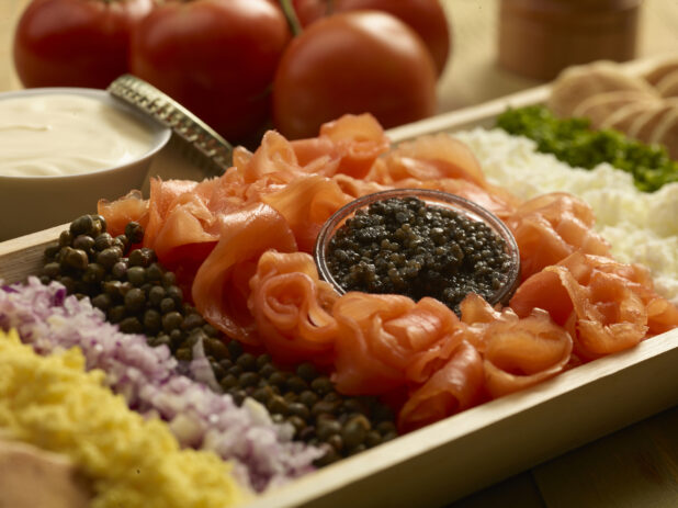Close up view of a smoked salmon platter with red onions, capers, caviar, in a rectangular wood catering tray with cream cheese an whole tomatoes in the background
