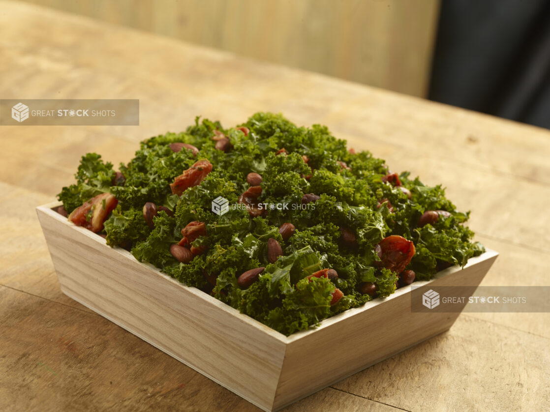 Kale salad with sun-dried tomatoes and whole almonds in a wood catering bowl on a wood table