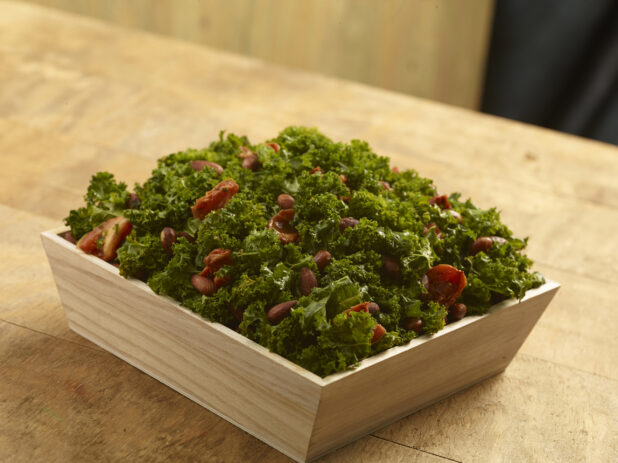 Kale salad with sun-dried tomatoes and whole almonds in a wood catering bowl on a wood table