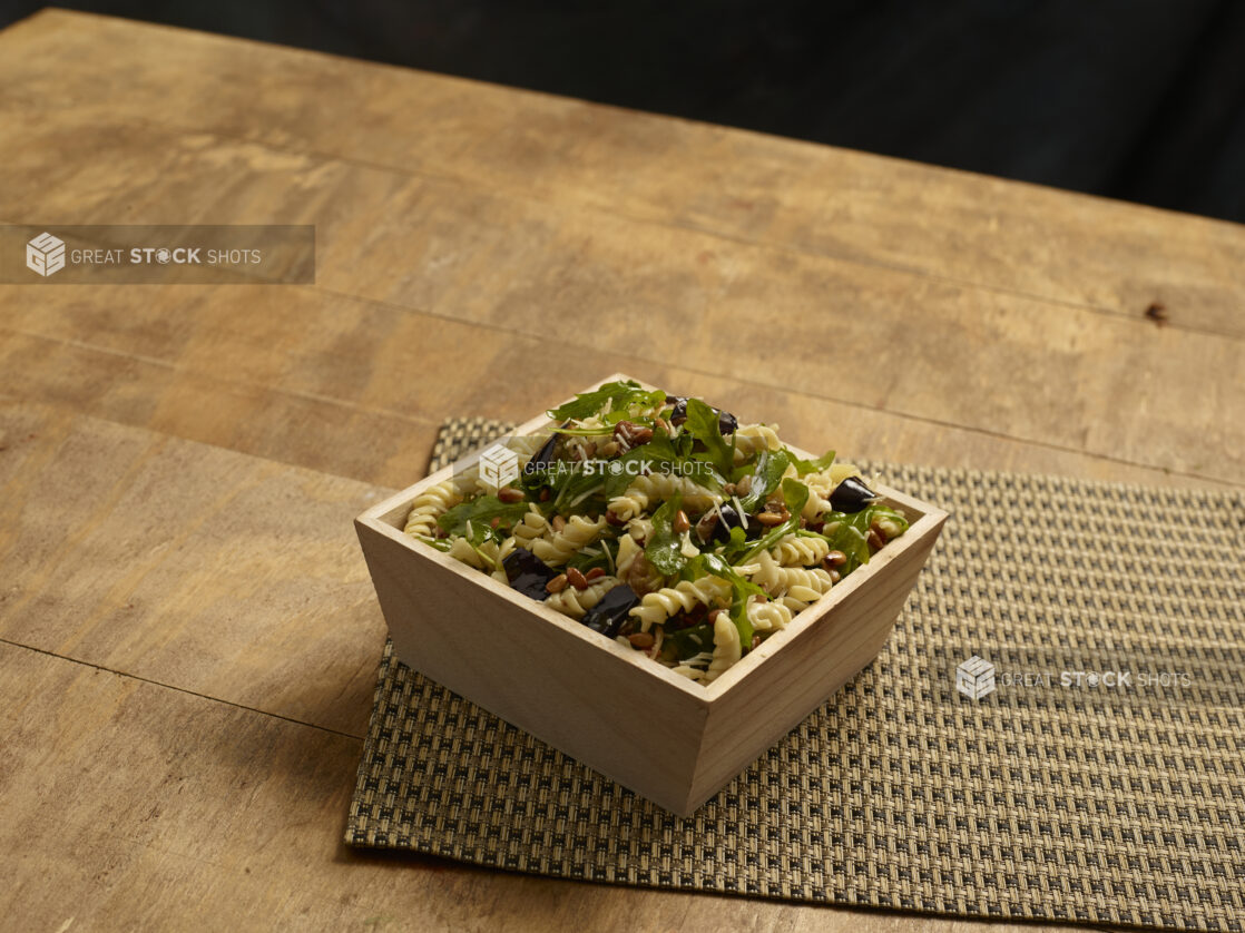 Vegetarian fusilli pasta salad with eggplant, roasted pine nuts and arugula in a wooden catering box on a wooden background