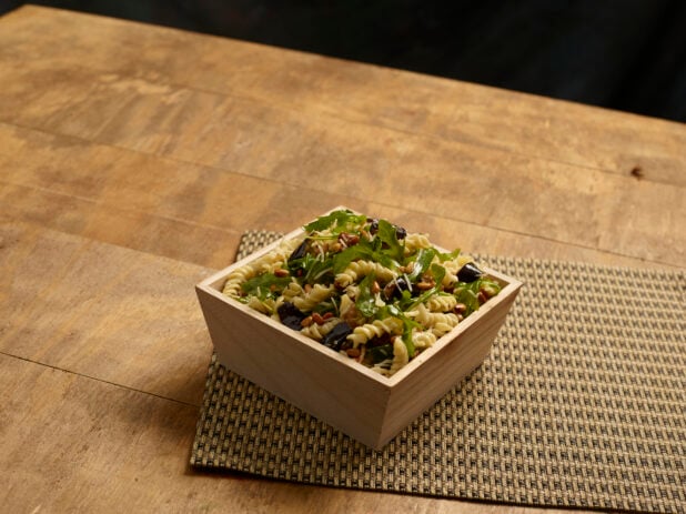 Vegetarian fusilli pasta salad with eggplant, roasted pine nuts and arugula in a wooden catering box on a wooden background