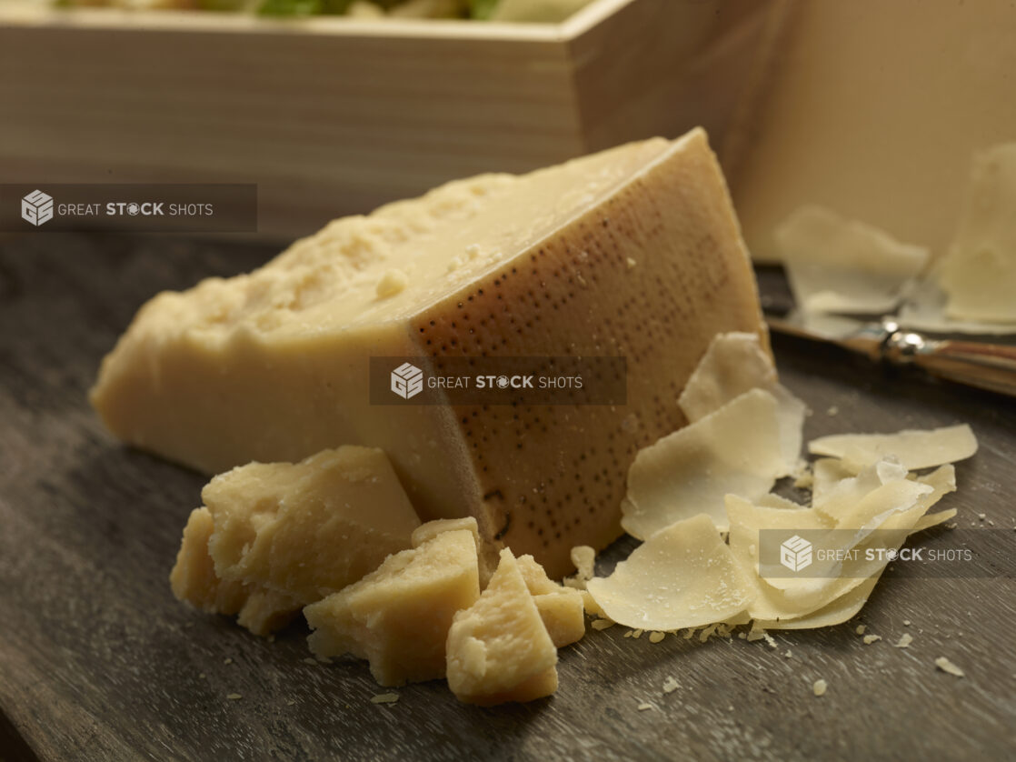 Whole block of parmesan cheese with chunks and shavings of parmesan on the side on a dark, close-up on wooden background