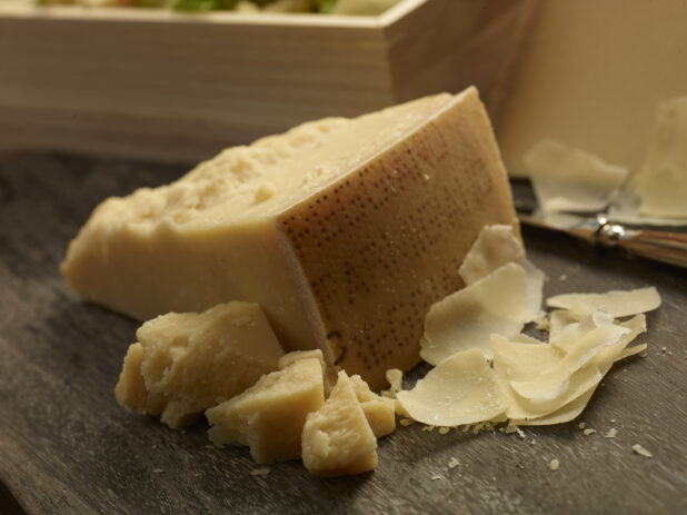 Whole block of parmesan cheese with chunks and shavings of parmesan on the side on a dark, close-up on wooden background