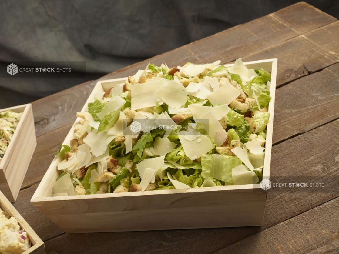 Caesar salad in a large wood catering box with croutons and shaved parmesan on a wooden background