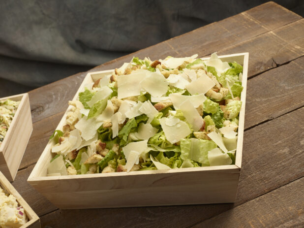 Caesar salad in a large wood catering box with croutons and shaved parmesan on a wooden background