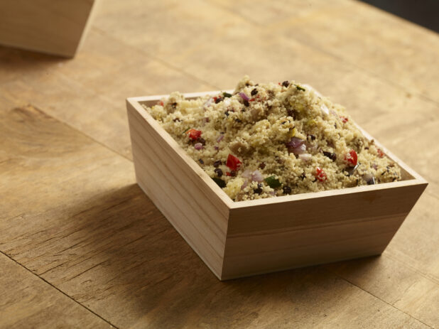 Cous cous salad with red bell peppers, red onion and currants in a square wood catering box on a wooden background