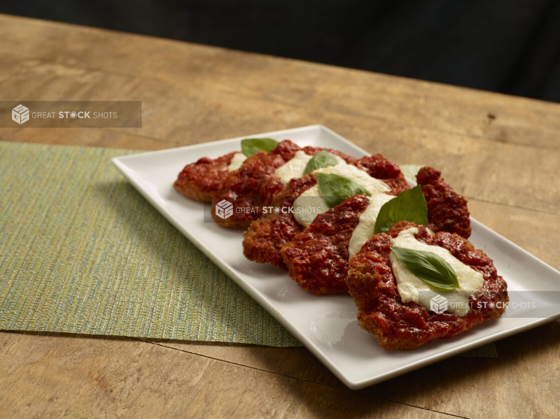 Pieces of chicken parmesan arranged a white ceramic platter, garnished with fresh basil