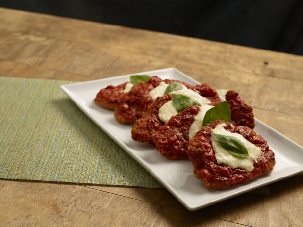 Pieces of chicken parmesan arranged a white ceramic platter, garnished with fresh basil