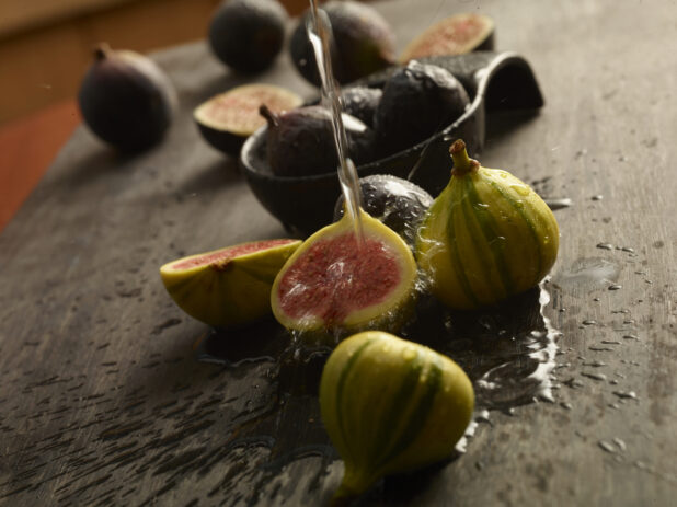 Bunch of fresh figs on a wood table with some cut in half with water being poured onto one