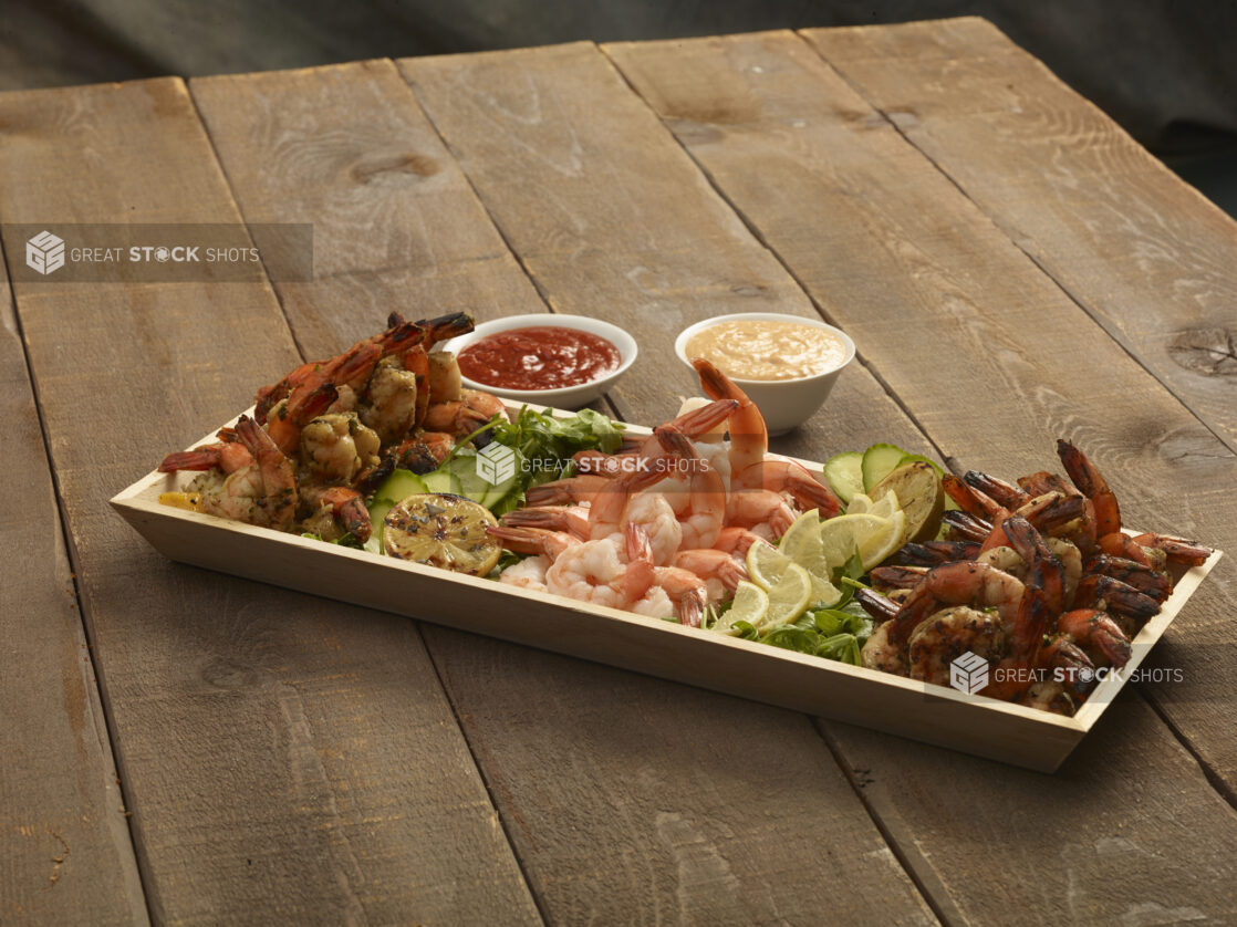 Shrimp appetizer platter with grilled lemon and lemon slices on a rectangular wood catering tray with seafood sauce and chipotle mayo on the side on a wooden background