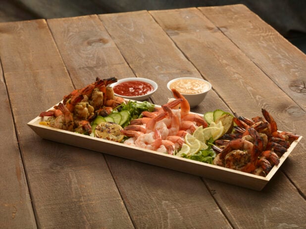 Shrimp appetizer platter with grilled lemon and lemon slices on a rectangular wood catering tray with seafood sauce and chipotle mayo on the side on a wooden background