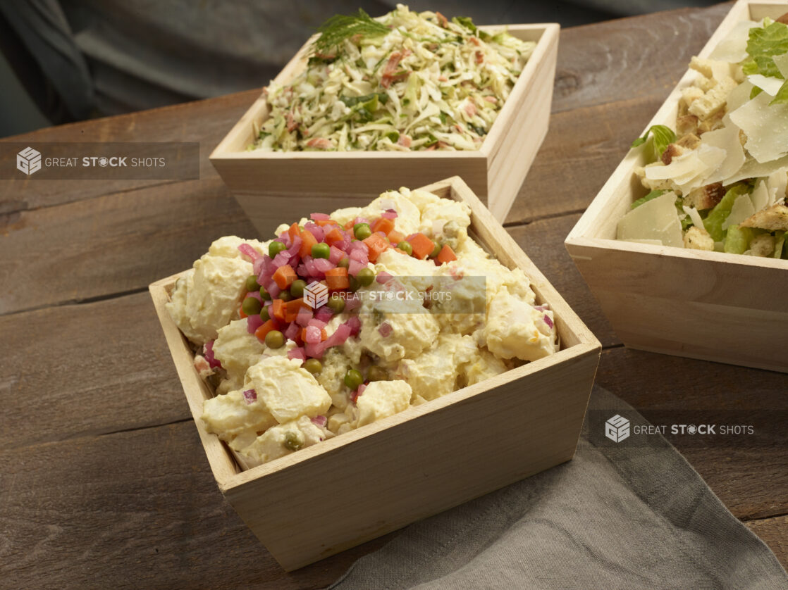 Potato salad, coleslaw and Caesar salad all in square wooden catering boxes on a wooden background