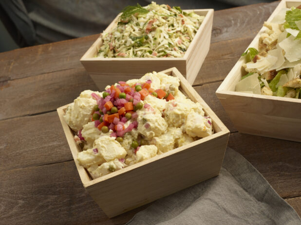 Potato salad, coleslaw and Caesar salad all in square wooden catering boxes on a wooden background