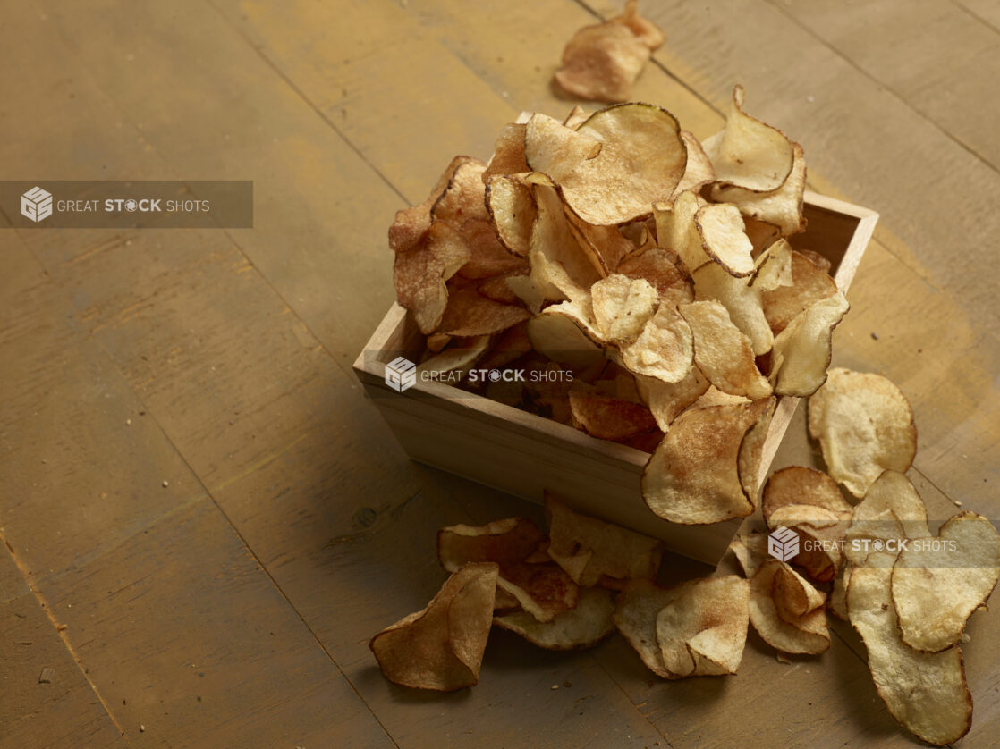 Rustic potato chips in and around a square wooden catering box, on a wooden background