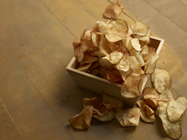 Rustic potato chips in and around a square wooden catering box, on a wooden background