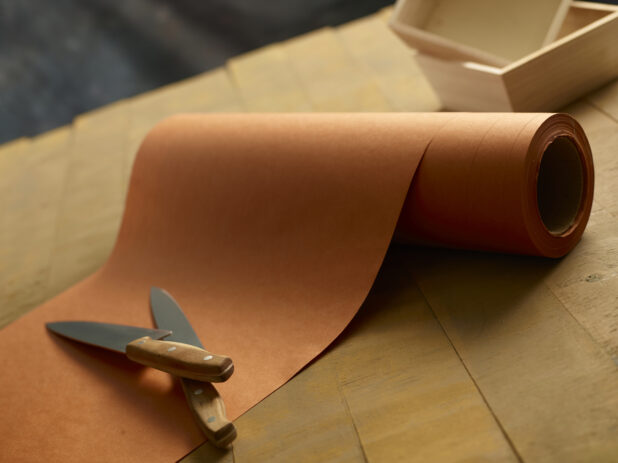 A roll of butchers paper, with 2 chefs knives and 2 small wood catering boxes in the background on a wooden table