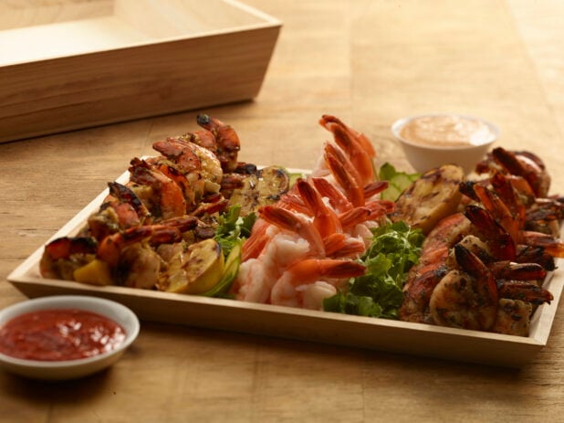 Shrimp appetizer platter on a rectangular wood catering tray with grilled lemons, seafood sauce and chipotle dipping sauce on a wooden background
