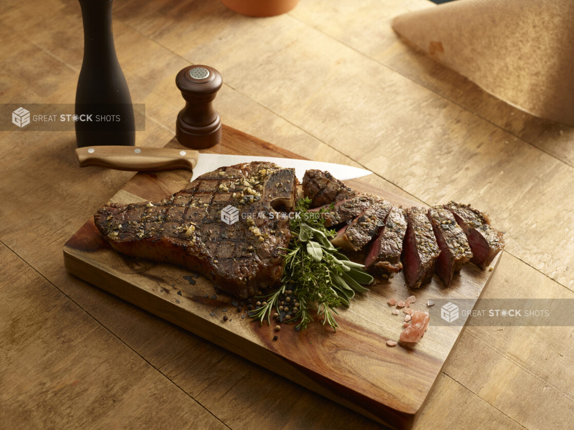 Grilled porterhouse steak, with slices of steak, on a wood board with a fresh herb garnish and chef knife, salt shaker and pepper grinder on a wooden background