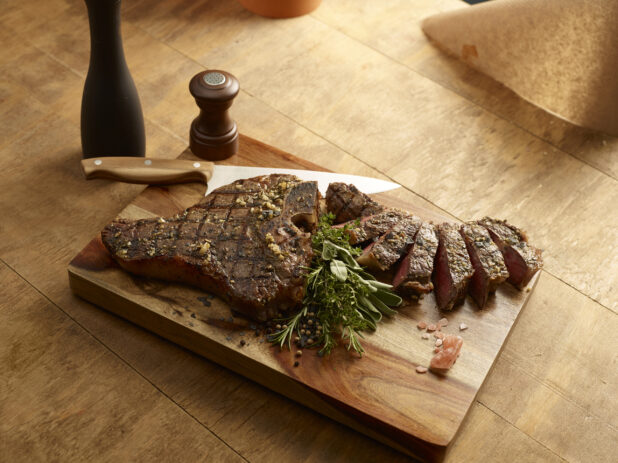 Grilled porterhouse steak, with slices of steak, on a wood board with a fresh herb garnish and chef knife, salt shaker and pepper grinder on a wooden background