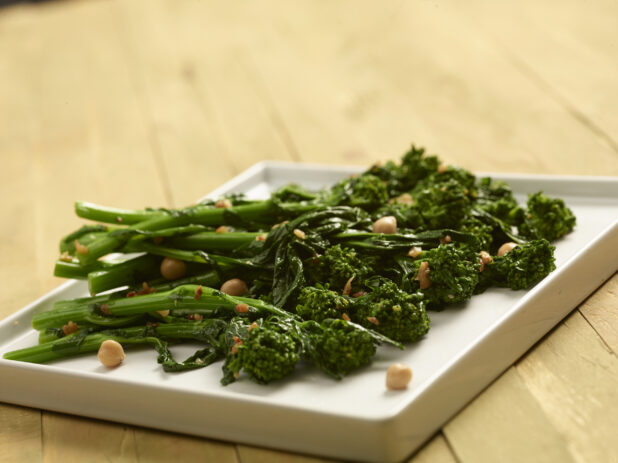 Steamed rapini with garlic and chick peas on a rectangular white plate on a wooden background, close-up