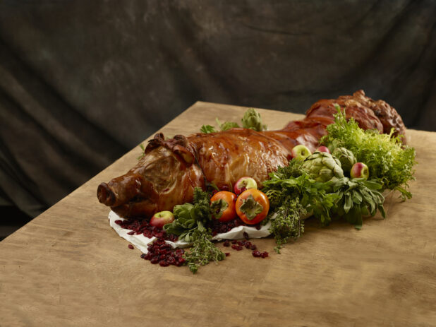 Whole roasted suckling pig with fresh apples, artichoke, persimmons, dried cranberries and fresh herbs surrounding all on a wooden table