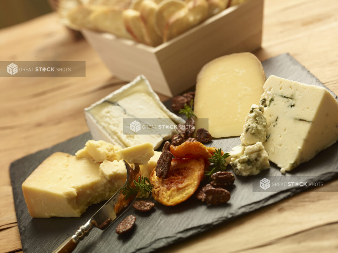 Cheese tray with various cheeses, dried fruit and candied pecans on a slate platter with assorted breads in a wood catering box in the background