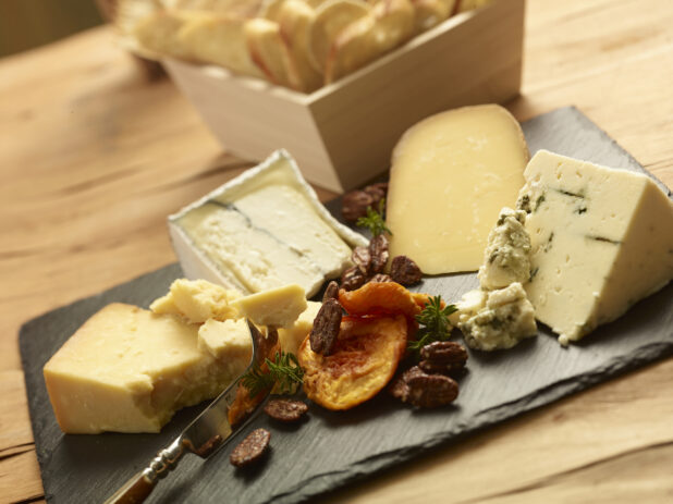Cheese tray with various cheeses, dried fruit and candied pecans on a slate platter with assorted breads in a wood catering box in the background