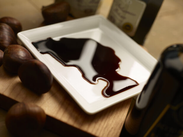 Balsamic vinegar on a small white rectangular plate with vinegar bottles and whole chestnuts surrounding