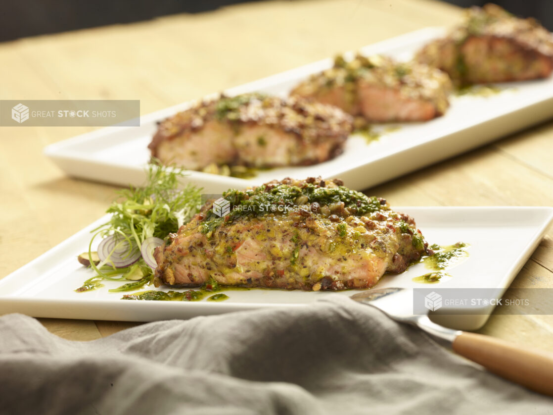 Grilled salmon on a rectangular with a pesto drizzle and fresh herb garnish with a platter of grilled salmon on a white platter in the background