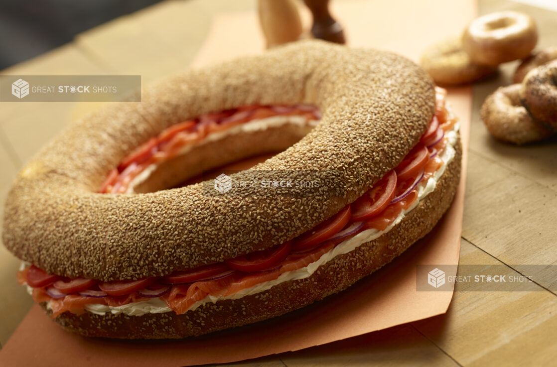 Giant sesame bagel with smoked salmon, cream cheese, tomatoes and red onion with whole uncut bagels in the background