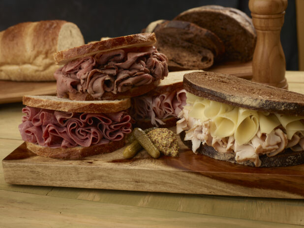 Deli sandwiches, corned beef, roast beef, ham, and turkey with swiss cheese on light and dark rye with pickles and grainy mustard on a wooden board with loaves of rye bread in the background