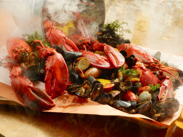 Metal bucket dumping out steaming hot seafood boil on to butcher paper in a tray on a wood table