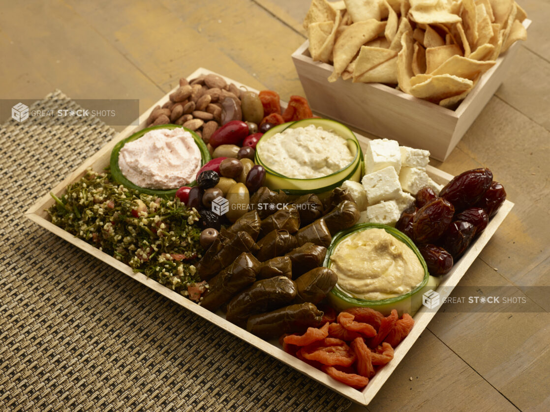 Mediterranean platter of stuffed grape leaves, tabouli, dates, dried apricots, feta cheese, olives and nuts with hummus, baba ganouj and taramasalata on a wood catering tray with a square catering box with pita chips