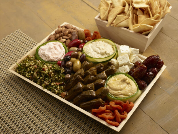 Mediterranean platter of stuffed grape leaves, tabouli, dates, dried apricots, feta cheese, olives and nuts with hummus, baba ganouj and taramasalata on a wood catering tray with a square catering box with pita chips