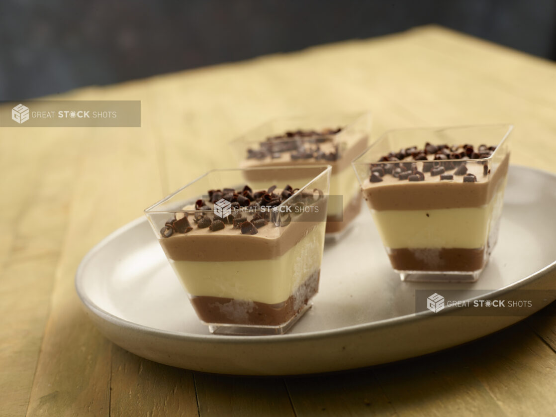 Triple chocolate mousse in square clear ramekins with chocolate shavings on top on a earth tone ceramic plate on a wooden background