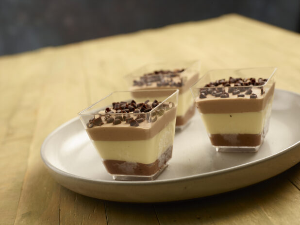 Triple chocolate mousse in square clear ramekins with chocolate shavings on top on a earth tone ceramic plate on a wooden background