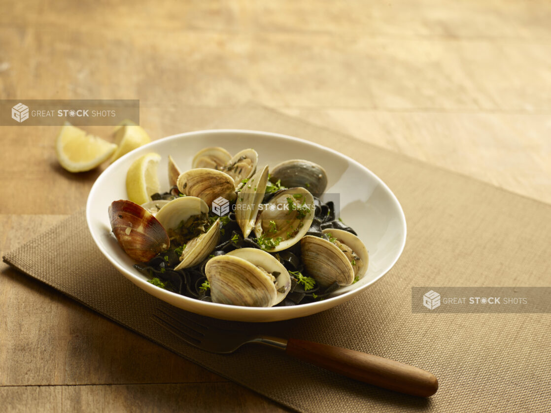 Squid ink linguine with clams and herbs in a white bowl with lemon wedges on a wood table