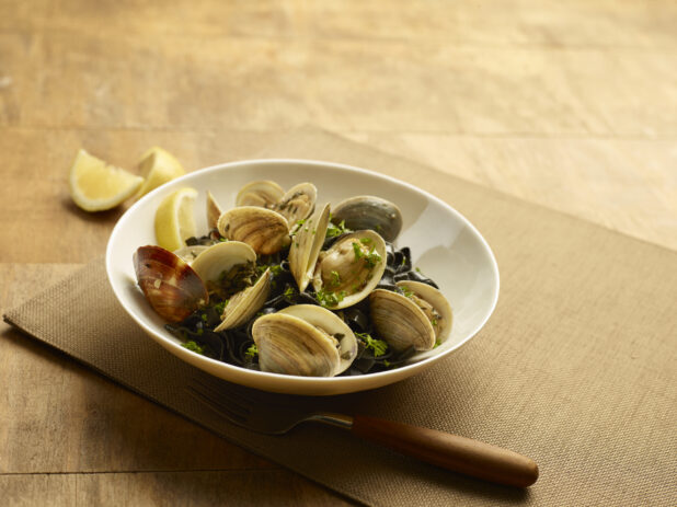 Squid ink linguine with clams and herbs in a white bowl with lemon wedges on a wood table